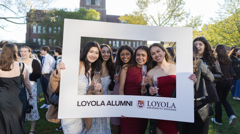 Five alumnae in formal dress enjoy drinks at an alumni event. They hold a frame for the photo that says Loyola Alumni.